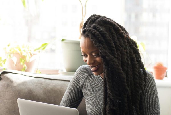 Black woman sitting on a sofa, smiling while working on a laptop indoors.