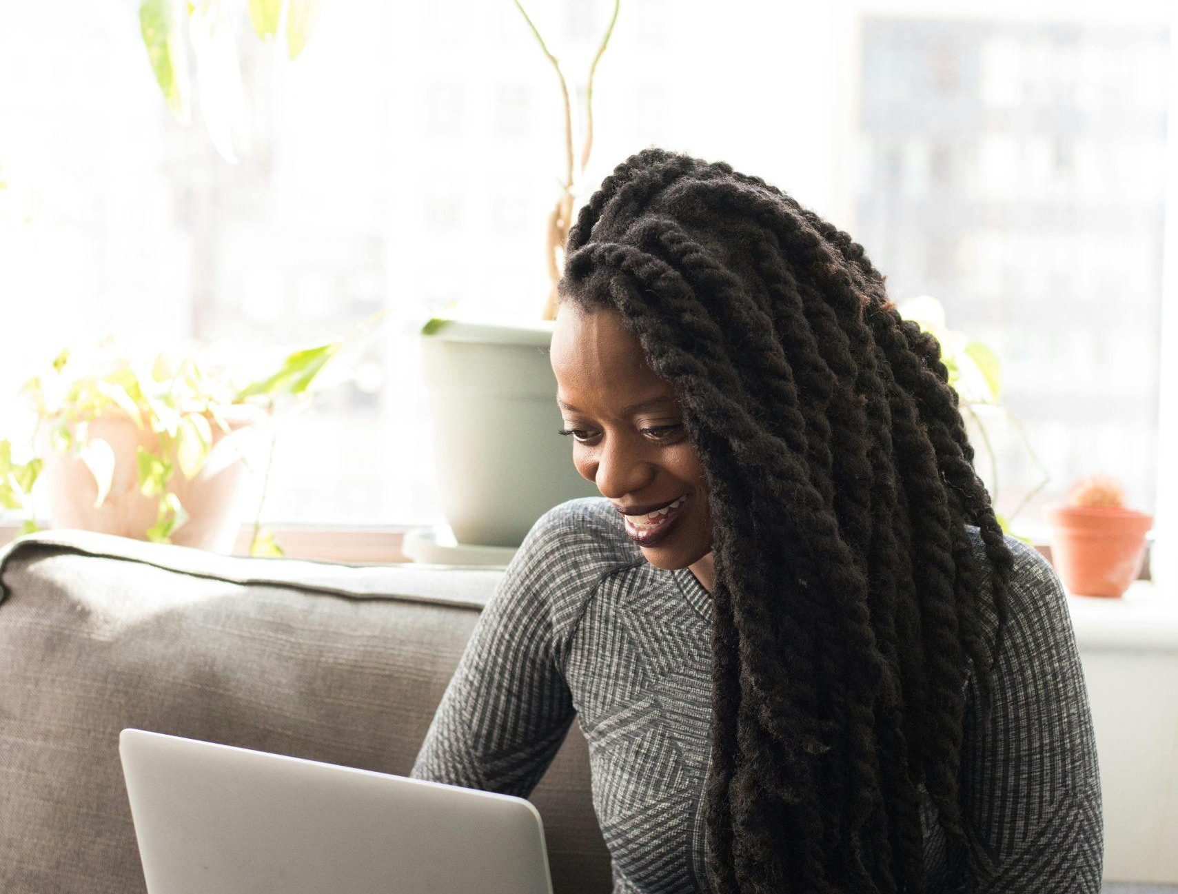 Black woman sitting on a sofa, smiling while working on a laptop indoors.
